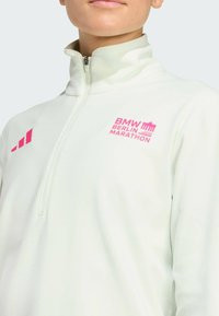 Person iført en hvid fleece med kvart lynlås og rosa BMW Berlin Marathon-logo på brystet samt et tre-stribet design på skulderen.