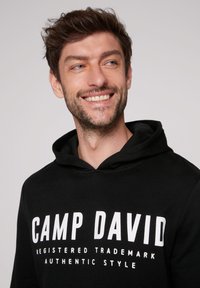 Sudadera negra con una gran impresión blanca que dice "CAMP DAVID" y texto más pequeño debajo, material de algodón con una textura suave, diseño tipo pullover.