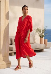 Femme en robe midi rouge avec détail froncé marchant près d'un canapé beige et d'une plante en pot sur une terrasse ensoleillée au bord de la mer.