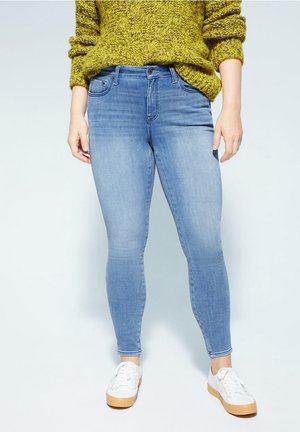 Person iført lyseblå skinny jeans, hvide sneakers og en grøn strikket sweater, stående mod en ensfarvet baggrund.