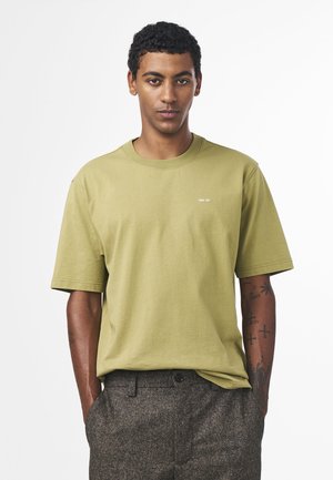 Olivengrøn bomulds t-shirt med rund hals og korte ærmer; lille hvidt logo på brystet. Modellen har brune strukturerede bukser på.