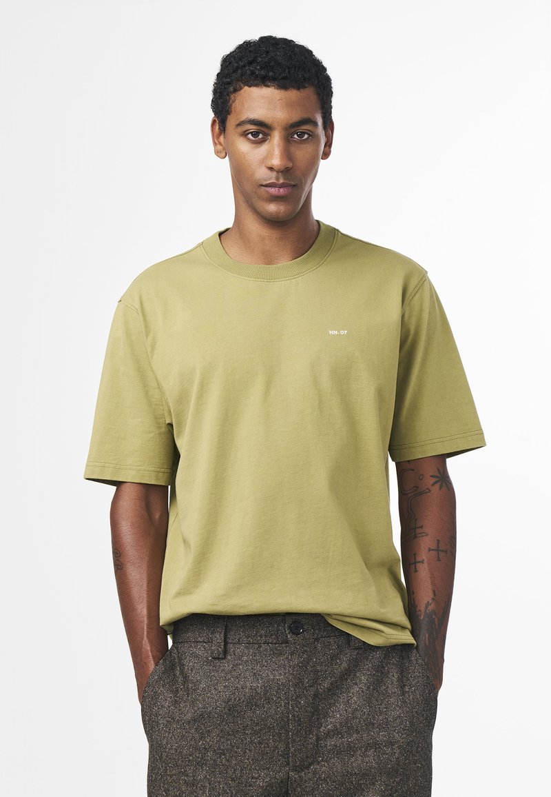 Olivgrön bomullst-shirt med rund hals och korta ärmar; liten vit logotyp på bröstet. Modellen har på sig bruna strukturerade byxor.
