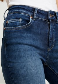 Jeans de denim azul oscuro con una textura suave, que presentan un corte ajustado, diseño de cinco bolsillos y acentos en tono cobre en los botones y remaches.