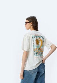 Biała, oversize t-shirt z kolorową grafiką przedstawiającą słońce i grzyby na plecach, z napisem "otwórz umysł" nad nią.
