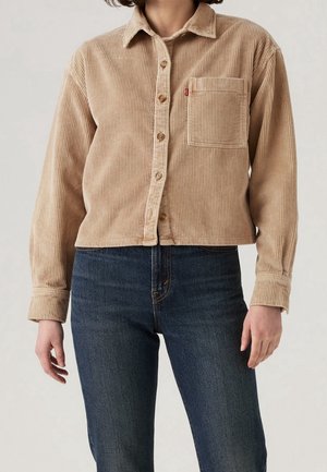 Person trägt beigefarbenes Cord-Hemd mit Brusttasche und dunkelblaue Jeans, dargestellt vom Hals bis zur Hüfte.