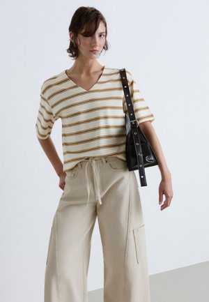 Young woman wearing a beige and white striped V-neck shirt, wide-leg beige pants, and carrying a black shoulder bag.