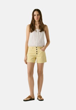 Donna in piedi che indossa una canotta bianca senza maniche, pantaloncini gialli a vita alta con bottoni e sandali marroni su uno sfondo neutro.