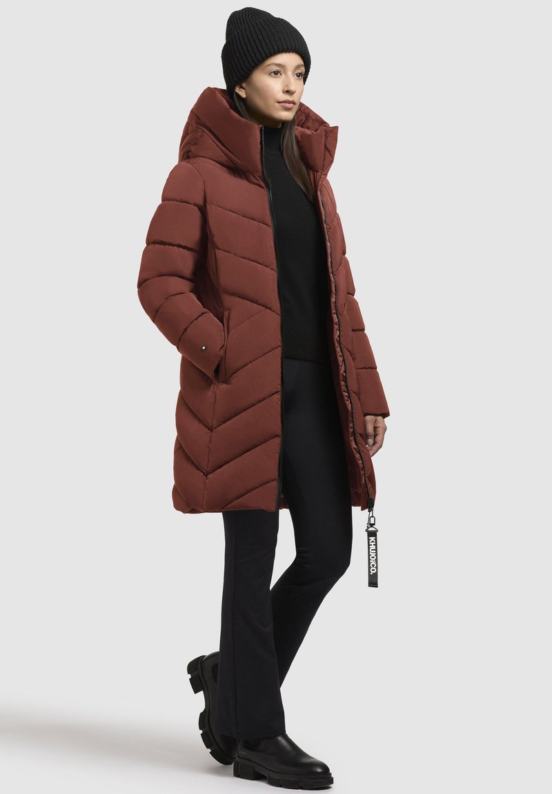 Giacca a vento di colore rosso scuro con colletto alto, cuciture a zig-zag, tasche laterali e chiusura con zip. Indossata sopra un maglione nero e pantaloni.