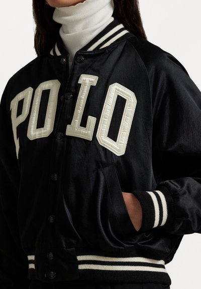 Mustast satiinist valmistatud bomber-jakk, millel on kreemivärvi tikandiga "POLO". Triibulised soonikkoes varrukate ja kaelaosa äärised. Includes küljetaskud.