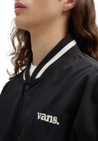 Osoba z długimi brązowymi włosami, ubrana w czarną kurtkę Vans z białymi paskami przy kołnierzu i haftowanym logo Vans na piersi.