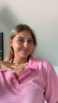 Polo shirt rose clair avec des rayures verticales blanches. Le col est de style classique et le tissu semble doux. Une personne sourit et fait un geste.