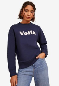 Tummansininen collegepaita, joka on valmistettu puuvillakankaasta ja jossa on pyöreä pääntie sekä ribbereunat. Edessä on vaalealla värillä brodeerattu "Voilà".