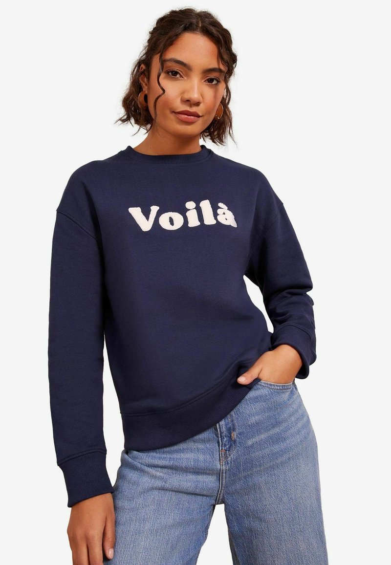 Tummansininen collegepaita, joka on valmistettu puuvillakankaasta ja jossa on pyöreä pääntie sekä ribbereunat. Edessä on vaalealla värillä brodeerattu "Voilà".