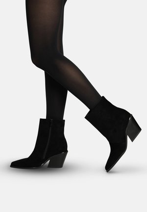Jambes en collants fins noirs portant des bottines en daim noir avec fermeture éclair latérale et talons carrés sur un fond clair.