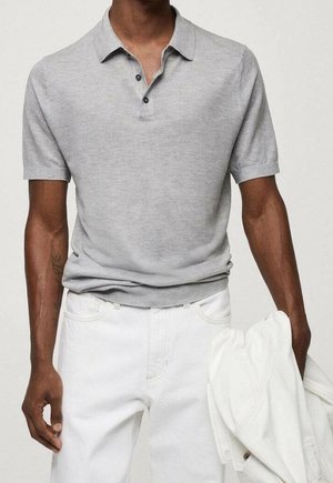 Homme portant un polo à manches courtes gris clair et un pantalon blanc, tenant une veste blanche pliée dans sa main gauche.