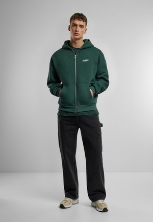 Dunkelgrüner Zip-Hoodie mit Kängurutasche, gerippten Bündchen und Saum; kombiniert mit schwarzen Hosen und beigen Sportschuhen.