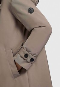 Manteau matelassé beige avec un bouton noir en accent sur le poignet gauche et une poche latérale. Texture lisse avec un design propre et moderne.