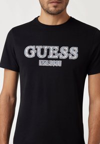 Juodas medvilnės marškinėliai su dideliu, grafišku "GUESS EST. 1981" spaudiniu baltos ir šviesiai mėlynos spalvų kombinacijoje ant krūtinės. Trumpomis rankovėmis ir apvaliu kaklu.