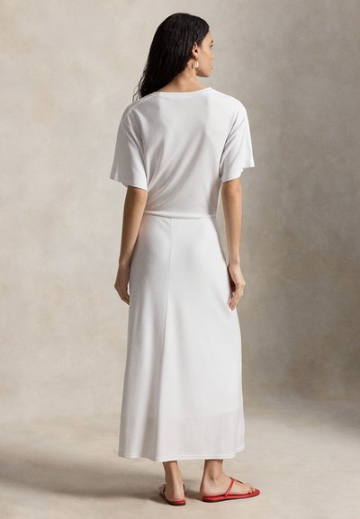 Γυναίκα με μακριά σκούρα μαλλιά που φοράει λευκό κοντομάνικο τοπ και μακριά φούστα με κόκκινα σανδάλια, στέκεται μπροστά σε απλό μπεζ φόντο.