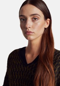 Sweter dzianinowy w ciemnozielonym kolorze z teksturowanym wzorem i czarnym kołnierzykiem w serek. Długie brązowe włosy opadają na ramie. Widoczna gładka skóra.