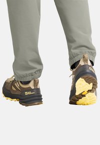 Chaussures de randonnée marron et grises avec des motifs texturés et des accents jaunes, dotées d'une semelle robuste et d'un talon rembourré, associées à un pantalon gris.