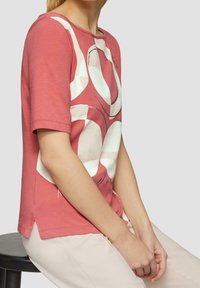 Rosa bomullströja med korta ärmar, med en vit abstrakt design och en sidöppning. Modellen sitter och bär ljusa byxor.