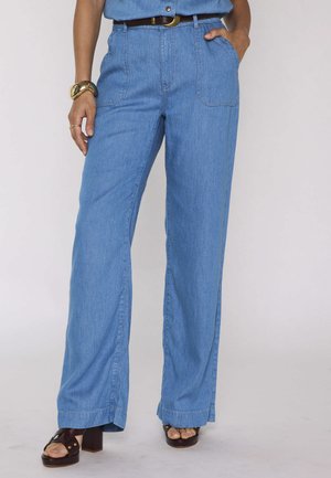 Vrouw draagt lichtblauwe high-waisted wijd vallende jeans met voorzakken, bruine riem, gouden armband en bruine sandalen met hak.