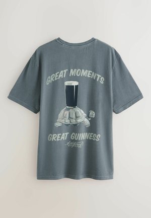 Oglekļa pelēks T-krekls ar uzrakstu "Lielie mirkļi Lielā Guinness", attēlojot bruņurupuci, kas nēsā melnā alus kausu ar putu uz muguras.