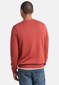 Trui in roestkleur, gemaakt van zachte stof. Ribgebreide manchetten en tailleband, eenvoudig ontwerp met ronde hals. Draag met blauwe spijkerbroek.
