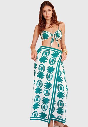 Mujer con un conjunto de dos piezas blanco y verde con estampado de piñas, que incluye un top corto anudado y una falda larga, acompañada de pulseras doradas.