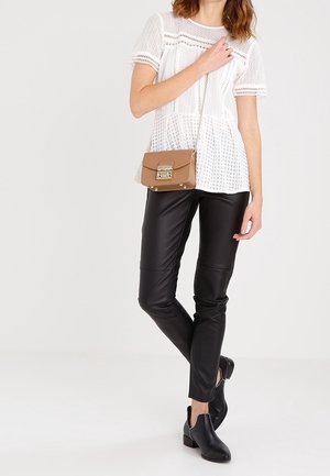 Nainen, joka pukeutuu valkoiseen pitsiseen lyhythihaiseen toppiin, mustiin nahkahousuihin, mustiin nilkkureihin ja kantaa pientä ruskeaa olkalaukkua, jossa on hopeinen solki.