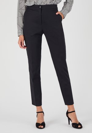 Pantalons noirs sur mesure avec une coupe ajustée, dotés d'une fermeture à bouton, de poches latérales et d'une texture lisse. Portés avec des sandales à talons hauts noires.