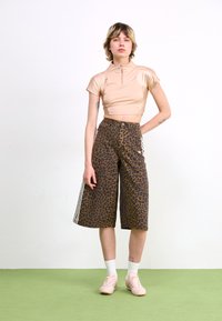 Young person wearing a beige cropped zip-up top, leopard print wide-leg shorts, white socks, and beige sneakers standing on green floor.