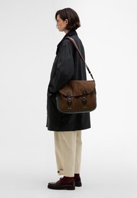 Καφέ καμβάς τσάντα messenger με μαύρες δερμάτινες λεπτομέρειες, ρυθμιζόμενο λουρί και δύο μπροστινές αγκράφες. Σε ένα άτομο που φοράει μαύρο παλτό και μπεζ παντελόνια.