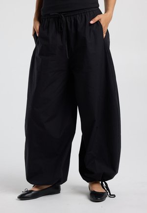 Personne portant un pantalon noir ample avec cordons de serrage aux ourlets et des ballerines noires, mains dans les poches, debout sur un sol blanc.