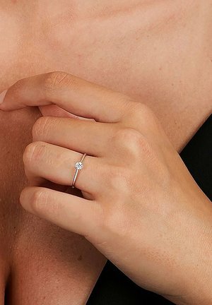 Silberner Ring mit einem einzelnen klaren Edelstein auf einem dünnen Band, getragen an einer Hand mit natürlichem Hautton vor einem dunklen Hintergrund.