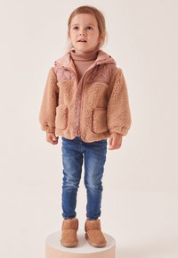 Veste en fausse fourrure marron avec des accents roses, fermeture éclair et deux poches frontales, associée à un jean bleu et des bottines marron. Enfant debout sur un socle.