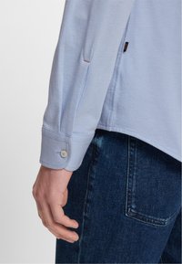 Camisa de manga larga azul claro con puños abotonados, que presenta una textura sutil y un detalle de abertura lateral, combinada con jeans azules oscuros.