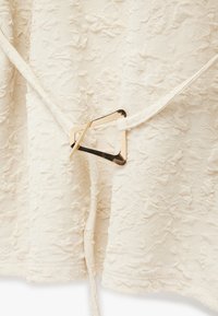 Cremefarvet tekstil med en guld, uregelmæssigt formet spænde, der fastgør to tekstilremme.