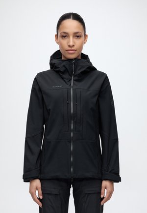 Une femme est face à l’objectif, vêtue d’une veste Mammut noire à capuche fermée par une fermeture éclair et d’un pantalon noir, sur un fond uni clair.