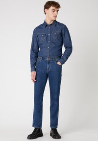 Denim overhemd met een puntkraag, twee borstzakken en contrasterende stiksels; gecombineerd met recht-toe-rechtdoor blauwe jeans en zwarte veterschoenen.