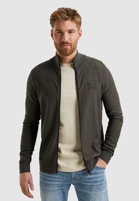 Temno zelena zip-up kardigan iz strukturiranega blaga, z visokim ovratnikom in aplikacijo na prsih, v kombinaciji z svetlo bež majico.