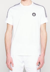 Man in witte korte mouwen Michael Kors-shirt en bijpassende broek met zwarte logopatch en merkstrepen op de schouders en zijkanten.