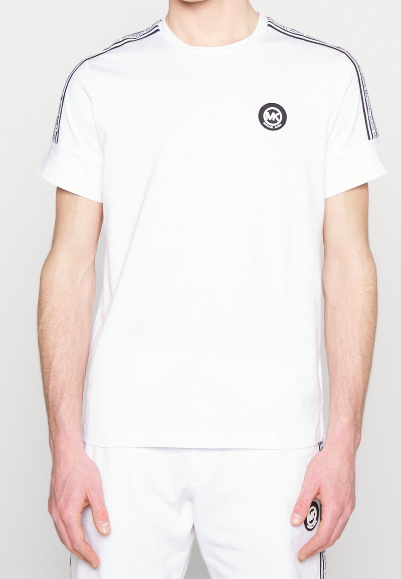 Man in witte korte mouwen Michael Kors-shirt en bijpassende broek met zwarte logopatch en merkstrepen op de schouders en zijkanten.