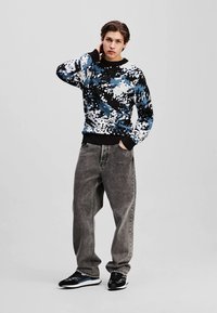 Sweater i sort, blå og hvid abstrakt mønster; elastiske manchetter. Grå slidte jeans; sorte sneakers med blå accenter.
