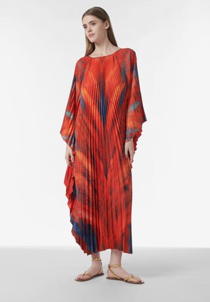 Mujer con un vestido rojo fluido y plisado con patrones azules y mangas sueltas, combinado con sandalias planas doradas, posando frente a un fondo blanco.