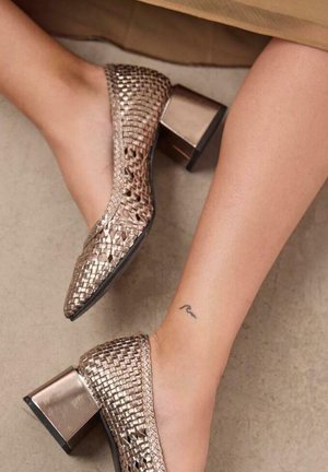 Zapatos de tacón de cuero trenzado color bronce con un acabado texturizado y tacones cuadrados. Accentos lisos y brillantes en la parte superior de los tacones.