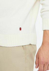 Bijela pletena pletiva s rebrastim rubom i manžetama. Ima mali crveni logotip na strani. Uparena s bež hlačama.