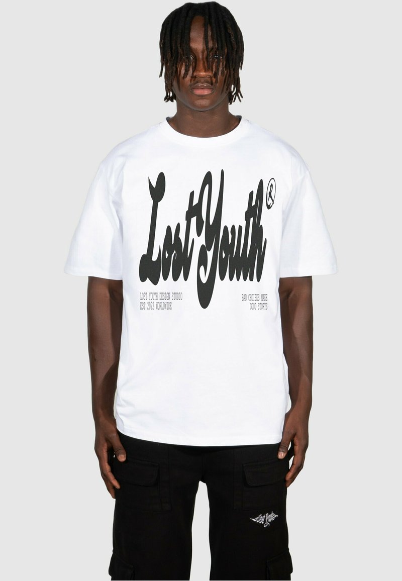 Vit bomull t-shirt med fet svart "Lost Youth" grafik och liten logotyp på bröstet, tillsammans med svarta cargobyxor med sidofickor.