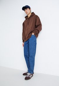 Brun hettegenser med kengurulomme, blå jeans og maroon treningssko med hvite striper. En caps fullfører det avslappede antrekket.